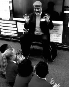 A black and white image of a man sitting on a chair in front of children sitting on the floor. 